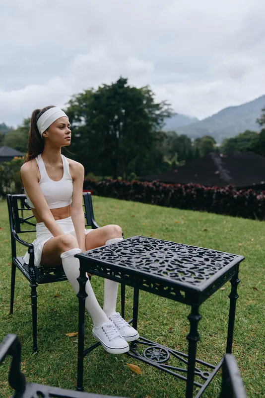 A young athletic woman in a white sports outfit sits on a chair, enjoying the serene outdoor scenery of a mountain landscape, reflecting calmness and confidence