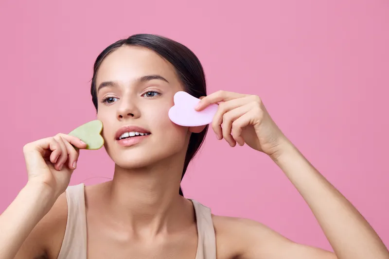 Young woman with a fresh, radiant smile using heart shaped facial tools against a pink background, showcasing a skincare routine