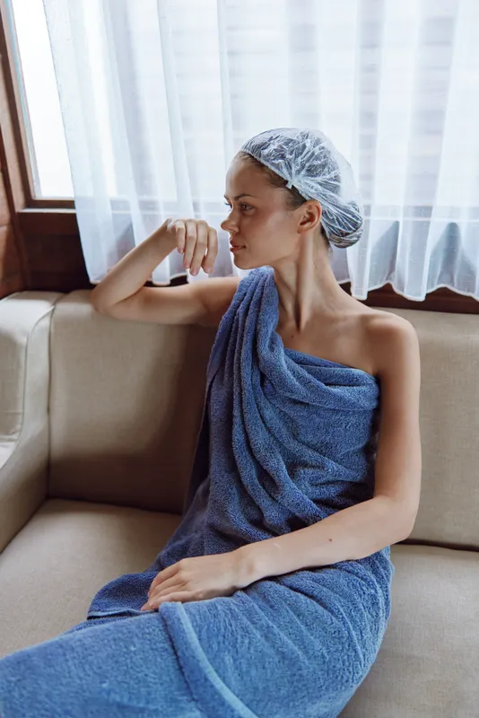 young woman wrapped in blue towel and wearing hair cap sitting thoughtfully on beige couch near window with sheer white curtains in natural light