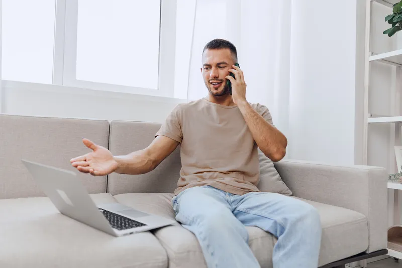 Engaged man in casual attire enjoys an honest phone conversation while connecting with his laptop, illustrating emotions related to modern work-life challenges.