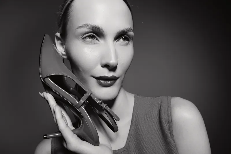 Portrait of white woman with pale skin holding high heel shoe near face. Studio black and white photo with fashion concept and elegant female model in close-up.