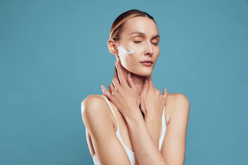 A young woman with clear skin applies cream on her face with closed eyes, touching her neck gently against a simple blue background. Concept for skincare and beauty care.