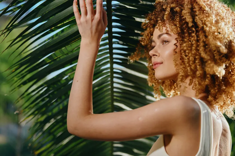 curly woman enjoying sunlight with lush green palm leaves background, natural beauty, warm summer day, outdoor portrait, relaxed mood