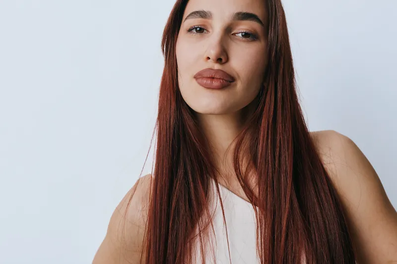 Young woman with long red hair and natural makeup posing against white background. Closeup portrait of confident female model with smooth skin and neutral expression in studio.