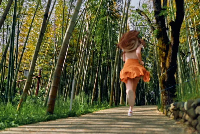 Woman running in orange dress along a sunlit path through a bamboo forest, motion blur and flowing hair convey freedom and adventure during a summer nature walk.