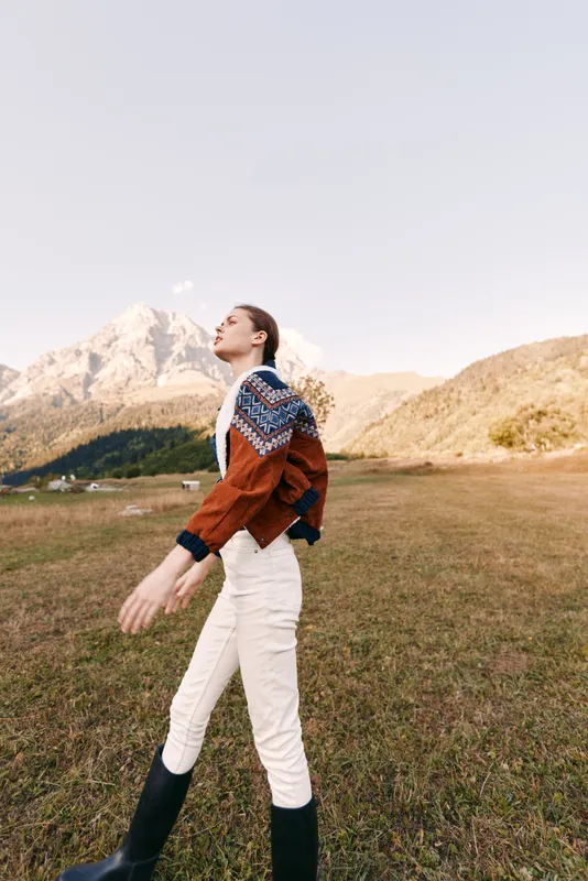 woman in patterned sweater walking across a grassy field with boots, mountain landscape and peaks in the background, autumn light and serene outdoor portrait conveying freedom and calm
