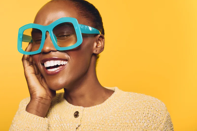Happy African American woman with dark skin wearing large blue glasses and yellow sweater smiles with closed eyes on yellow background. Concept of joy, fashion and beauty.