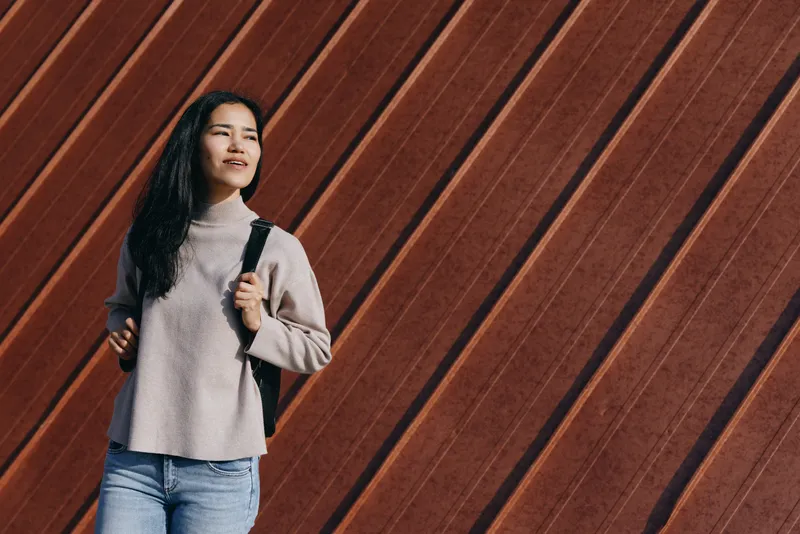 A woman with backpack, wearing a cozy sweater and jeans, stands beside diagonal wooden wall, embodying a casual outdoor moment, everyday style and urban street fashion, showcasing texture and natural