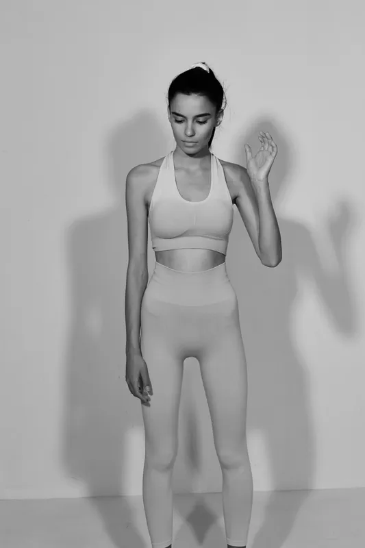 Black and white sport fitness woman wearing activewear, standing in a minimalistic studio with soft lighting, showing focused calm and body balance.