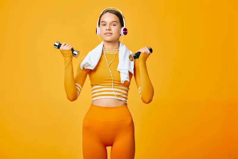 Fitness sport woman in bright orange outfit lifts dumbbells in a vibrant studio, showcasing workout routine, training focus and gym energy with towel and headphones.