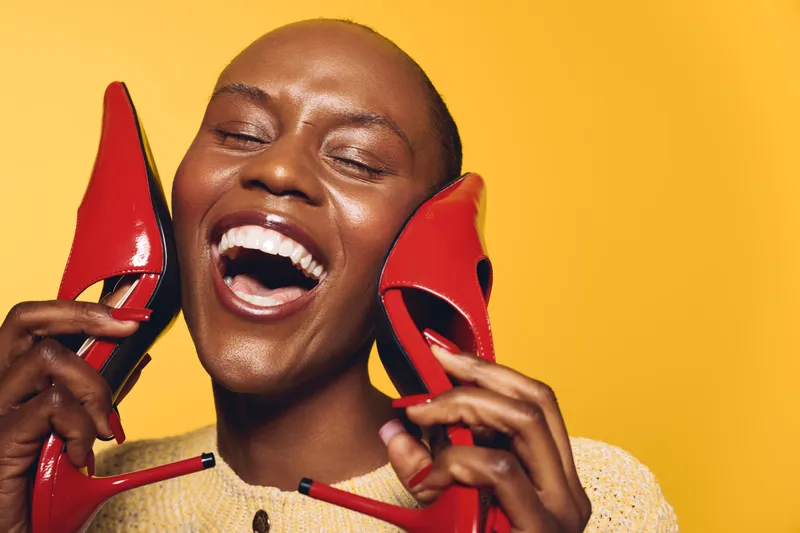 Happy black woman with dark skin laughs joyfully holding red high heels near her face on yellow background. Concept of fashion, beauty, and positive emotion in studio portrait.