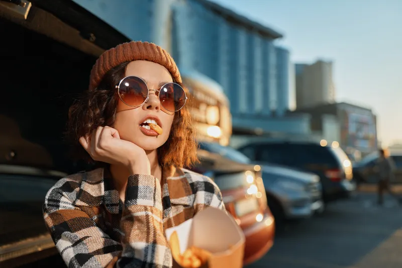 Woman in beanie and sunglasses eating fries by an open car trunk in a parking lot at golden hour, candid lifestyle portrait with authenticity, mindful living and emotional storytelling.