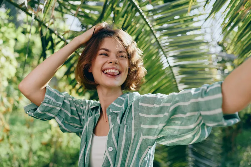 Joyful young woman with a wide smile stands under tropical palm fronds, wearing a green striped shirt, capturing a bright, carefree moment.