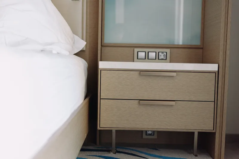 Bedside view with a wooden nightstand beside a white bed, clean lines and built in power outlets, creating a calm, modern bedroom look suitable for hotel rooms and contemporary homes.