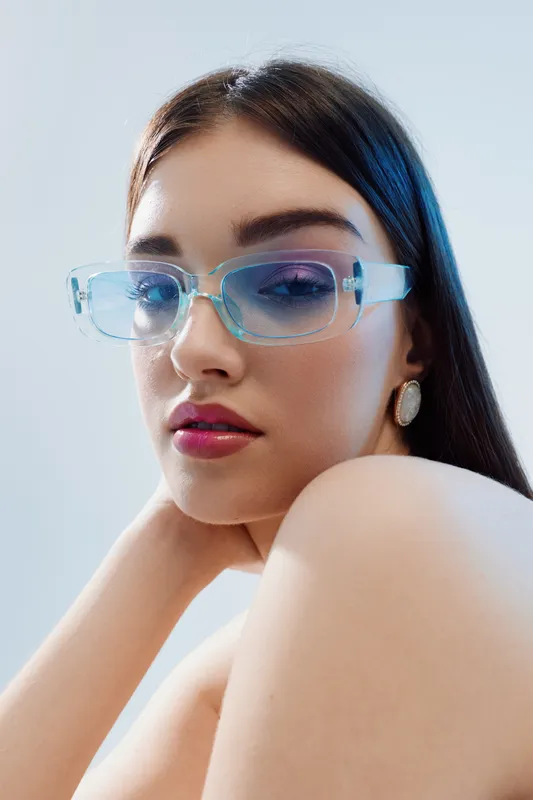 Young woman beauty fashion portrait with long dark hair wearing stylish transparent blue glasses and elegant earrings. Soft lighting highlights smooth skin and confident expression.