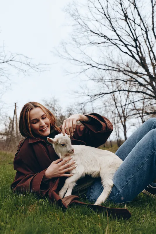 Joyful woman outdoors, sitting on the grass with a friendly goat kid, gentle touch, sunlit moment, casual jacket, relaxed posture, natural colors, tranquil countryside vibe, fresh breeze