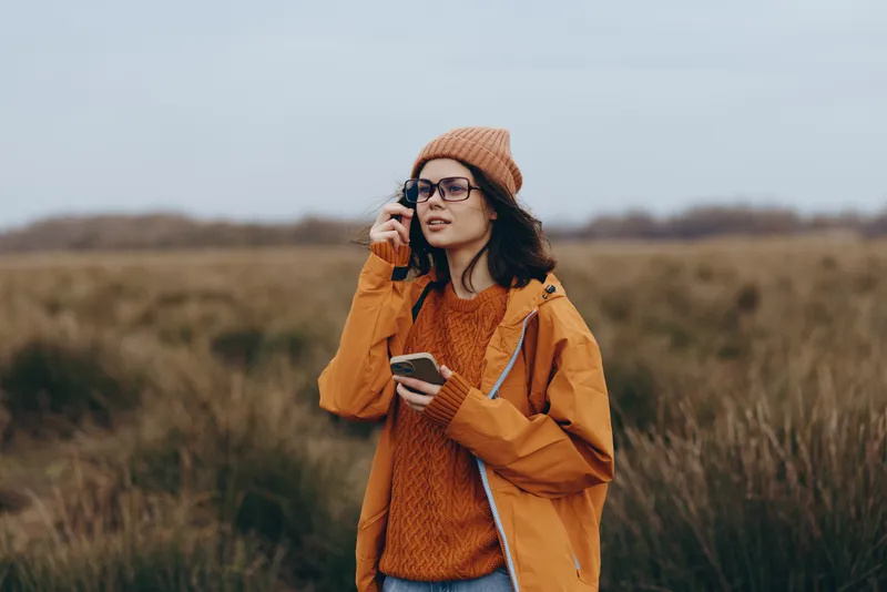 Woman lifestyle portrait outdoors in an autumn field, orange hat and sweater, holding a phone to her ear, speaking with a smile, rural meadow background