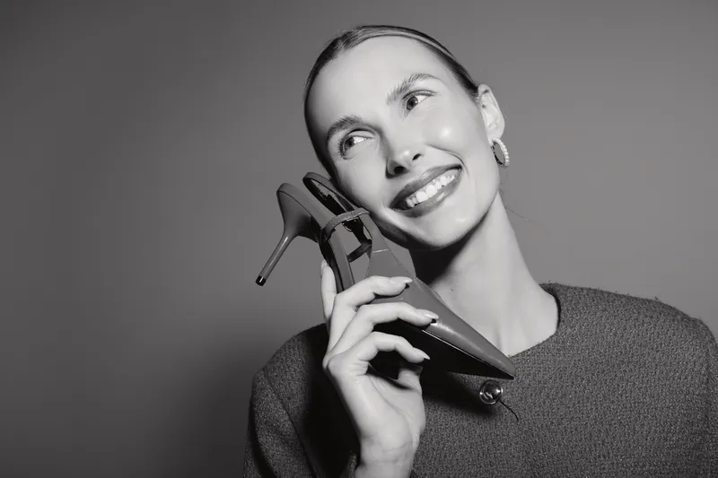 Smiling Caucasian woman with light skin holding a high heel shoe near her face in studio. Concept of fashion, style, and happiness.