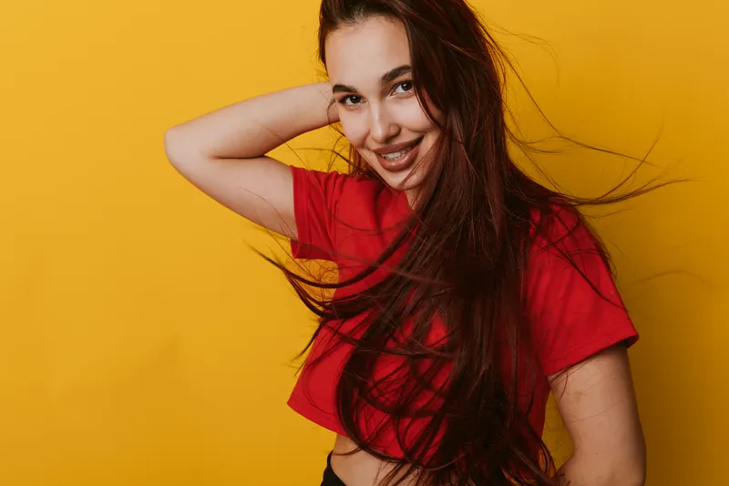 Young woman with long hair smiling and posing in red crop top against yellow background. Female model with carefree expression and natural beauty in casual outfit for portrait photography.