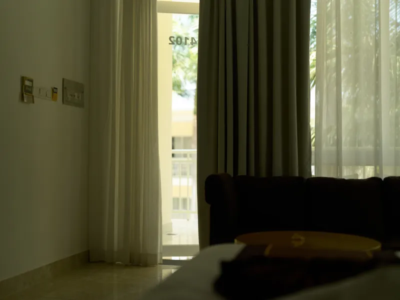 dimly lit hotel room interior with sheer curtains partially drawn, soft natural light filtering through window, cozy and quiet atmosphere