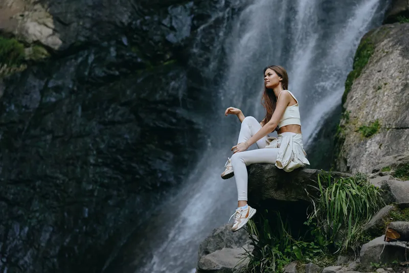young woman sitting by waterfall in casual white outfit, relaxed and enjoying nature, outdoors with rocky background and lush green plants, serene mood