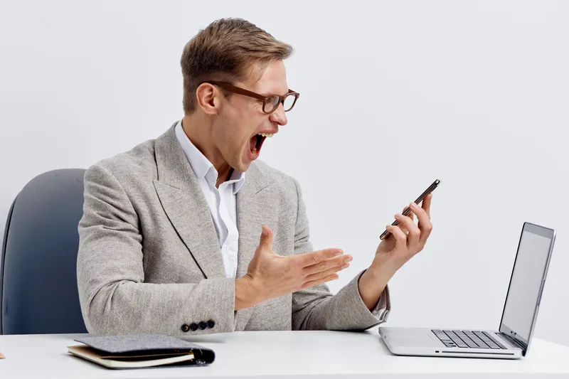 frustrated man using smartphone and laptop in business casual wear shouting in office digital nomad concept
