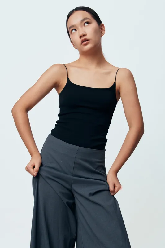 Young woman in black tank top and gray wide pants posing indoor with thoughtful expression. Fashion model with natural makeup and pulled back hair against plain white background.