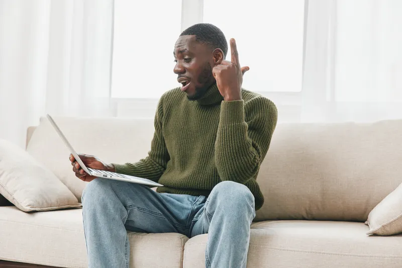 Young Black man engaged in an animated discussion on a laptop at home, wearing a green sweater and jeans, with a bright, cozy background emphasizing a modern indoor environment
