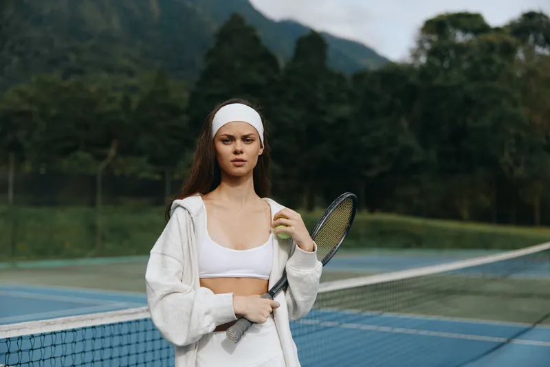 Young female tennis player in sportswear holding a racket with a confident expression on an outdoor court surrounded by greenery