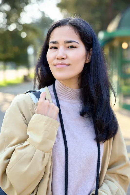Woman in a casual jacket with a bag stands outdoors in a park, sunlight filtering through trees, showcasing everyday fashion and a relaxed street style vibe.