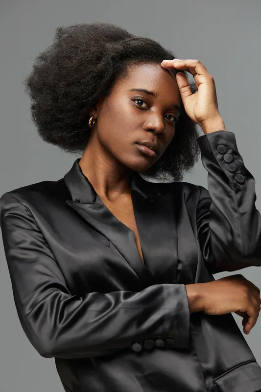 Elegant young woman with natural hair in a black satin blazer, exuding confidence and style against a neutral background