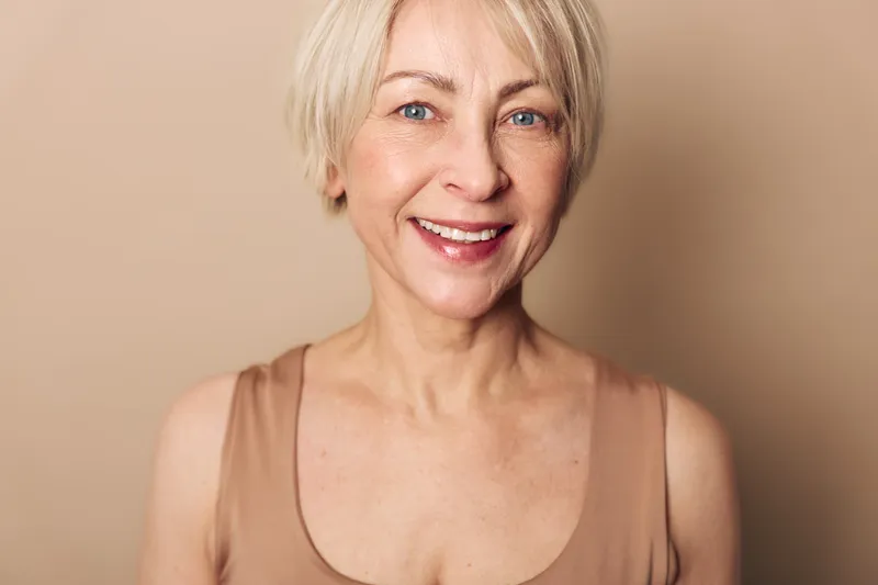 Smiling mature woman with short blonde hair wears beige top, looking at camera with natural makeup and clear blue eyes on soft warm background. Concept of beauty and confidence.