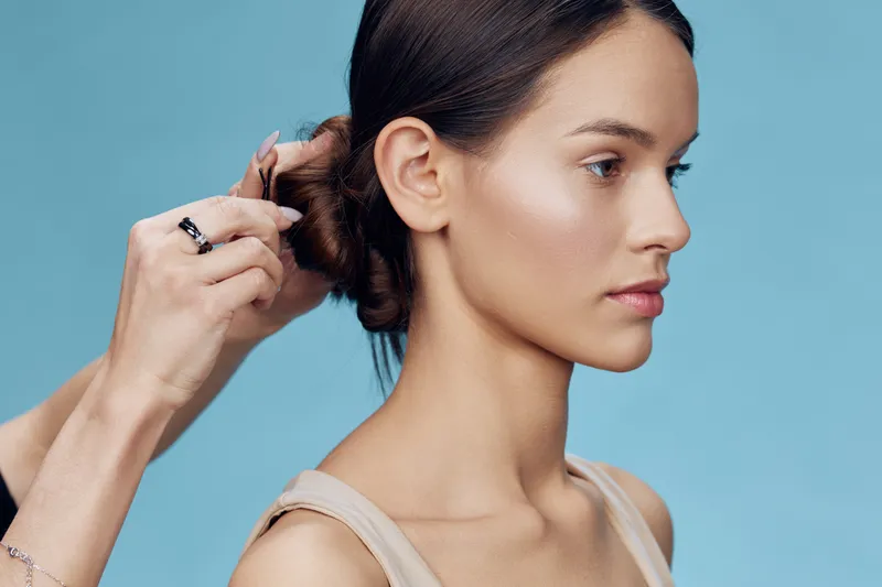 A beautiful young woman with a smooth, minimalistic hairstyle is having her hair styled against a soft blue background, emphasizing beauty and elegance