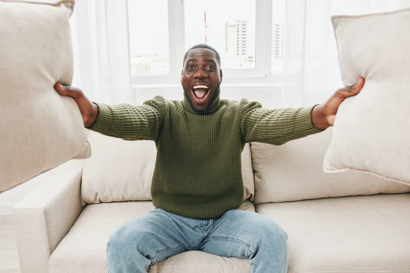 Happy African man celebrating at home with pillows, showcasing joy and excitement in a cozy living room setting with bright natural light