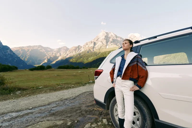 Woman leaning on white car by a mountain road, travel and nature landscape with panoramic valley. Stylish traveler in warm jacket standing outdoors for adventure and scenic drive.
