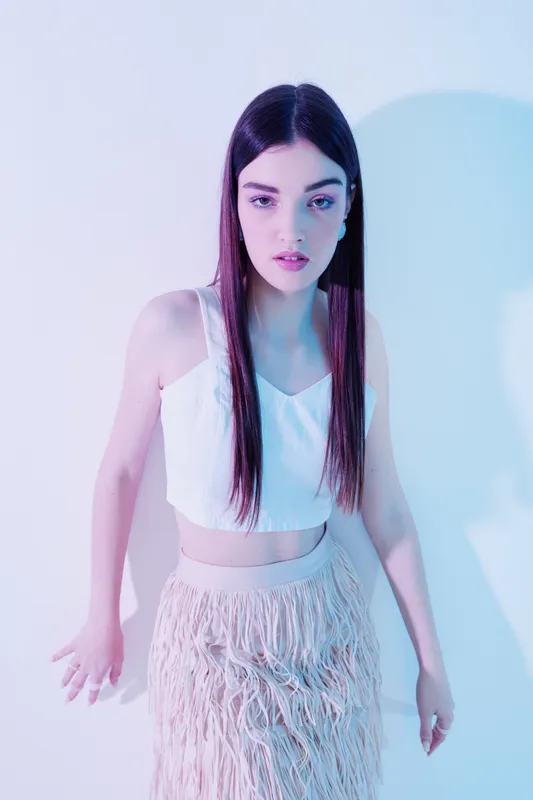 Young woman with long dark hair wearing a white crop top and fringe skirt poses in soft blue and pink studio lighting. Concept of beauty fashion and style.