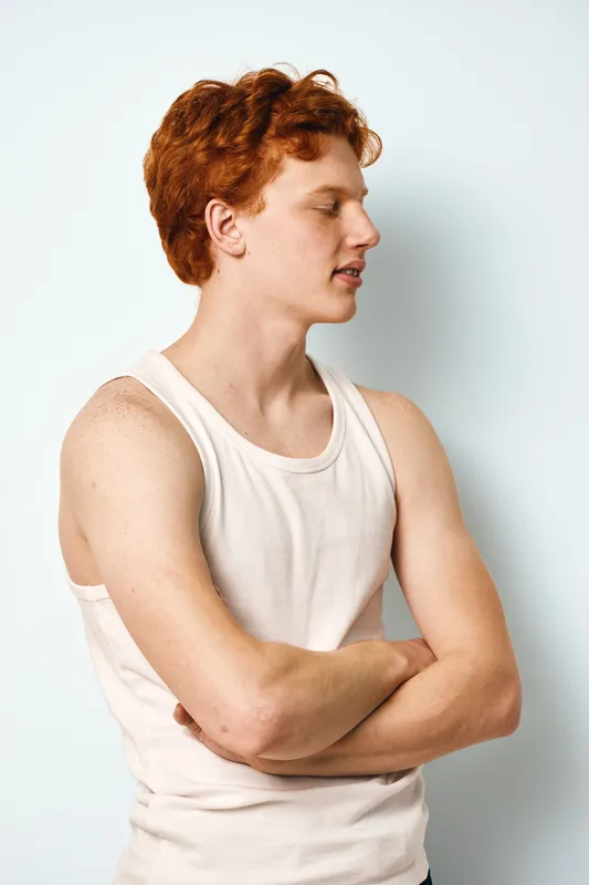 Young man with red hair in sleeveless shirt standing with crossed arms looking sideways on white background. Casual style and confident expression in studio portrait.