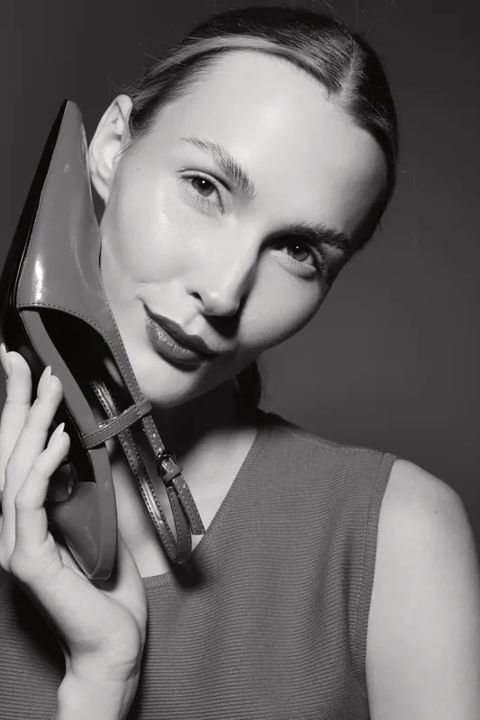 Closeup of a fair skinned Caucasian woman holding a shoe near her face, smiling softly, with smooth skin and slicked hair.