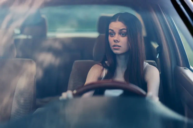 Young woman with long hair in a car, showing surprise and concern, wearing a casual outfit, captured in natural light, conveying emotion and tension.