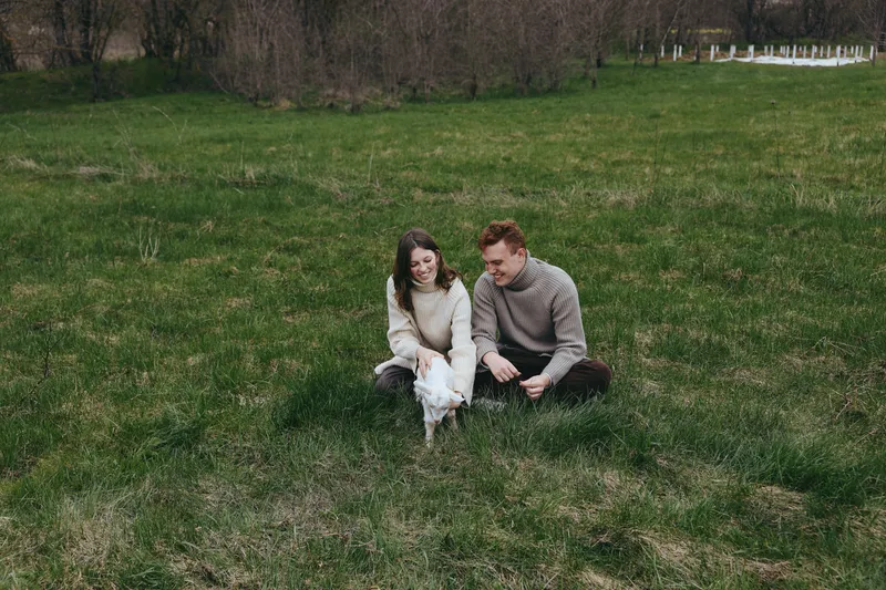 Cheerful couple outdoors with a playful goat kid, relaxing moment on a grassy field, soft sun, candid interaction, simple outfits, natural colors, tranquil countryside backdrop, warm connection,