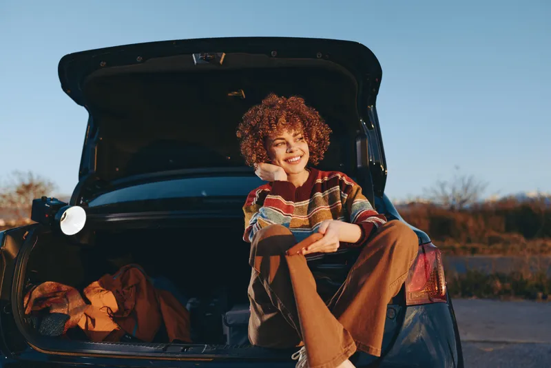 Girl child car trunk smile sunset outdoors lifestyle, sitting on open trunk at dusk, relaxed casual pose, warm golden light, with curly hair playful expression family road trip stop outdoors.