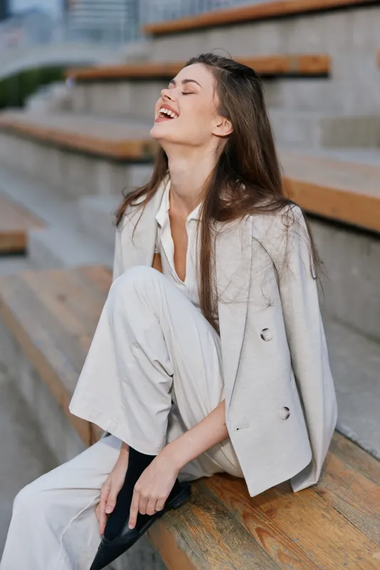 Young happy woman in light suit smiling joyfully seated on urban wooden stairs in casual fashion style autumn outfit