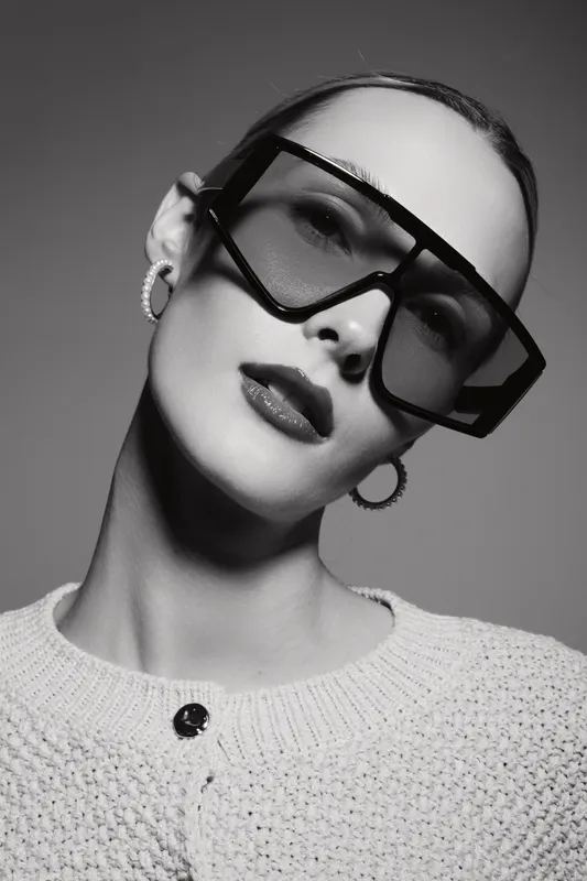 Portrait of a white woman with pale skin wearing oversized modern sunglasses and textured sweater. Studio shot with dramatic lighting, concept for fashion and style.