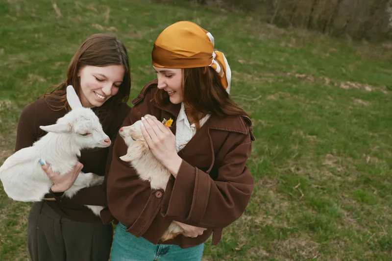 Two women smile while holding a white goat kid in a grassy field, capturing a warm lifestyle moment outdoors. Gentle, joyful interaction centers on animals and friendship.