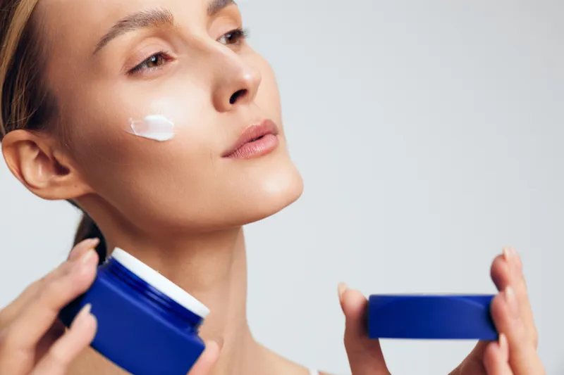 Young woman applies moisturizing cream on face holding blue jar skincare product with neutral expression isolated on light background for beauty and skin care concept.