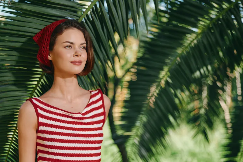 A stylish woman stands outdoors among lush tropical plants, wearing a red striped sleeveless top and a matching headscarf. She gazes forward with calm confidence.