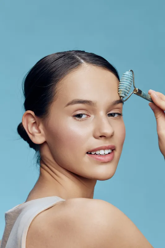 Young woman applying skincare with a facial roller, showcasing glowing skin against a light blue background, radiating freshness and beauty