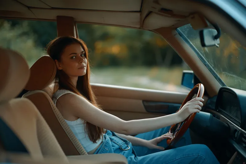 Young woman driving vintage car in natural setting, wearing casual outfit with denim overalls, showcasing a calm and confident expression.