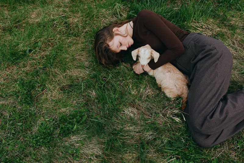 Woman lies on grass cuddling a small dog in a serene outdoor moment, soft light, casual clothing, peaceful mood, intimate lifestyle scene of companionship and nature.