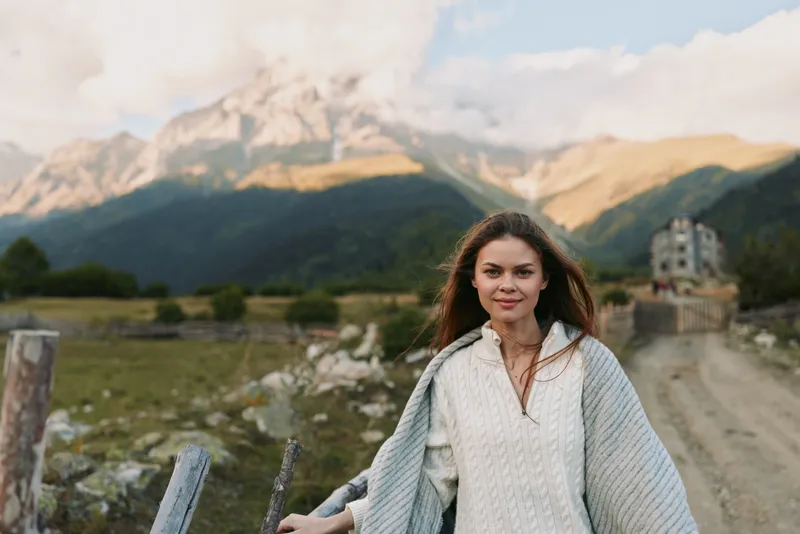 Young woman with long hair wearing a white sweater and shawl standing outdoors in a mountainous landscape. Concept of lifestyle, nature, and travel adventure.